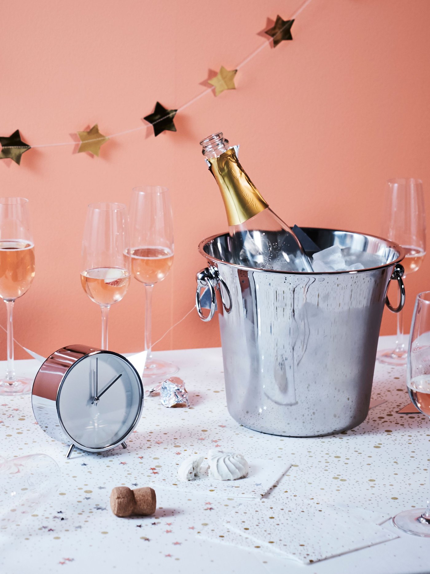Een zilverkleurige SOLDRÄNKT champagnekoeler met daarin een fles bubbels voor een roze achtergrond met wat STORSINT champagneglazen op tafel.