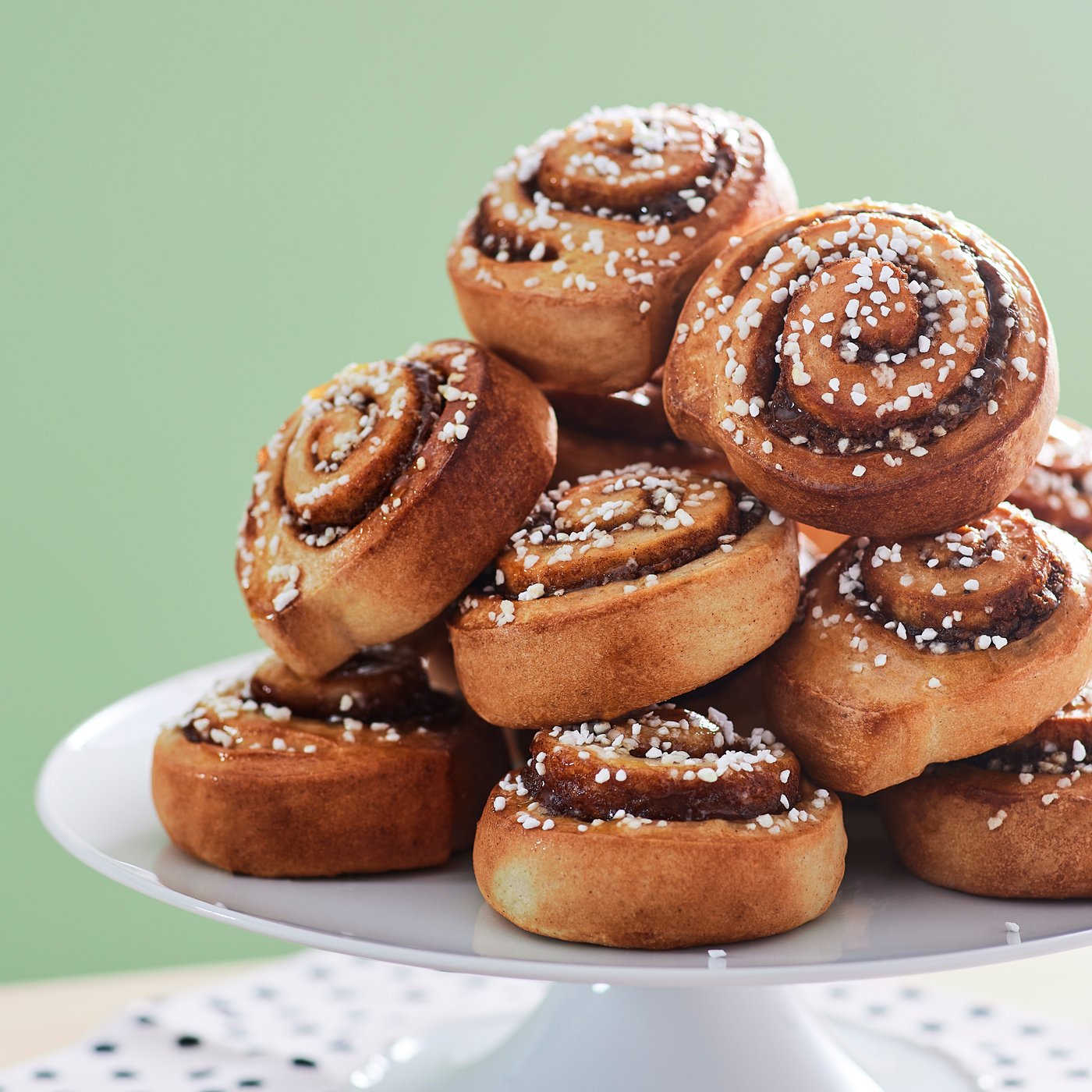 Rollos de canela IKEA con azúcar espolvoreada encima montados uno sobre otro arriba de una base blanca, atrás una pared verde.