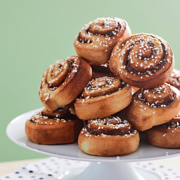 Rollos de canela IKEA con azúcar espolvoreada encima montados uno sobre otro arriba de una base blanca, atrás una pared verde.
