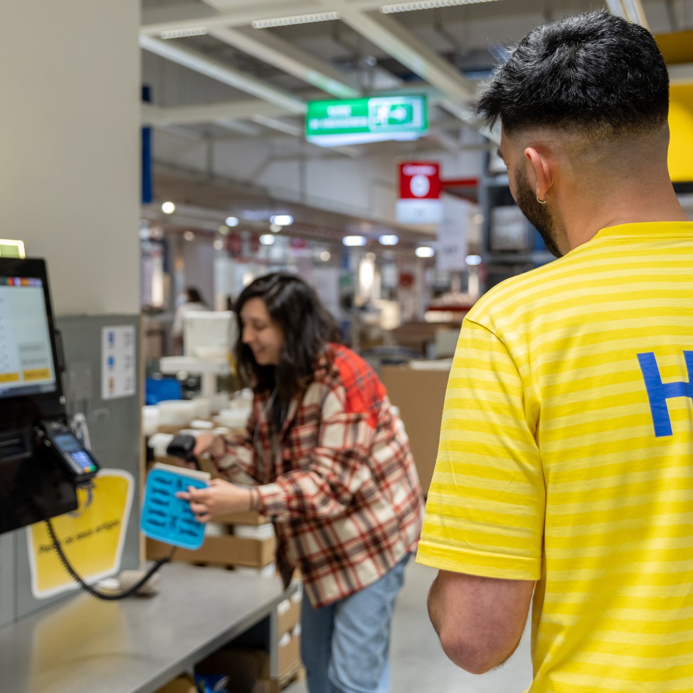 En kund klädd i rutig skjorta betalar sina varor i en självbetjäningskassa. I förgrunden syns en IKEA medarbetare med arbetströja med ordet Hej på ryggen på sig.