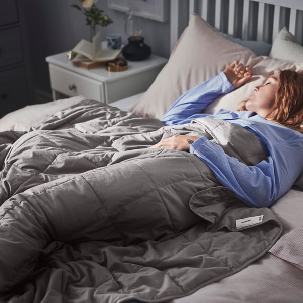 Una mujer durmiendo sobre una cama tapada con un cobrelecho ODONVIDE y al costado una mesita de noche TYSSEDAL.