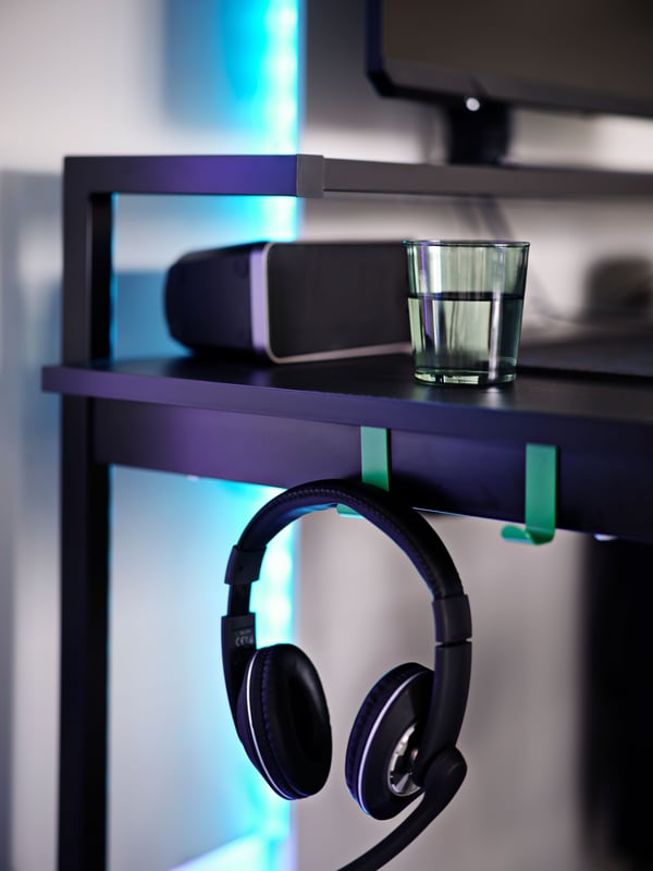 A headset hangs on one of two green metal hooks attached to the side below a black UTVISNING gaming desk with shelf.