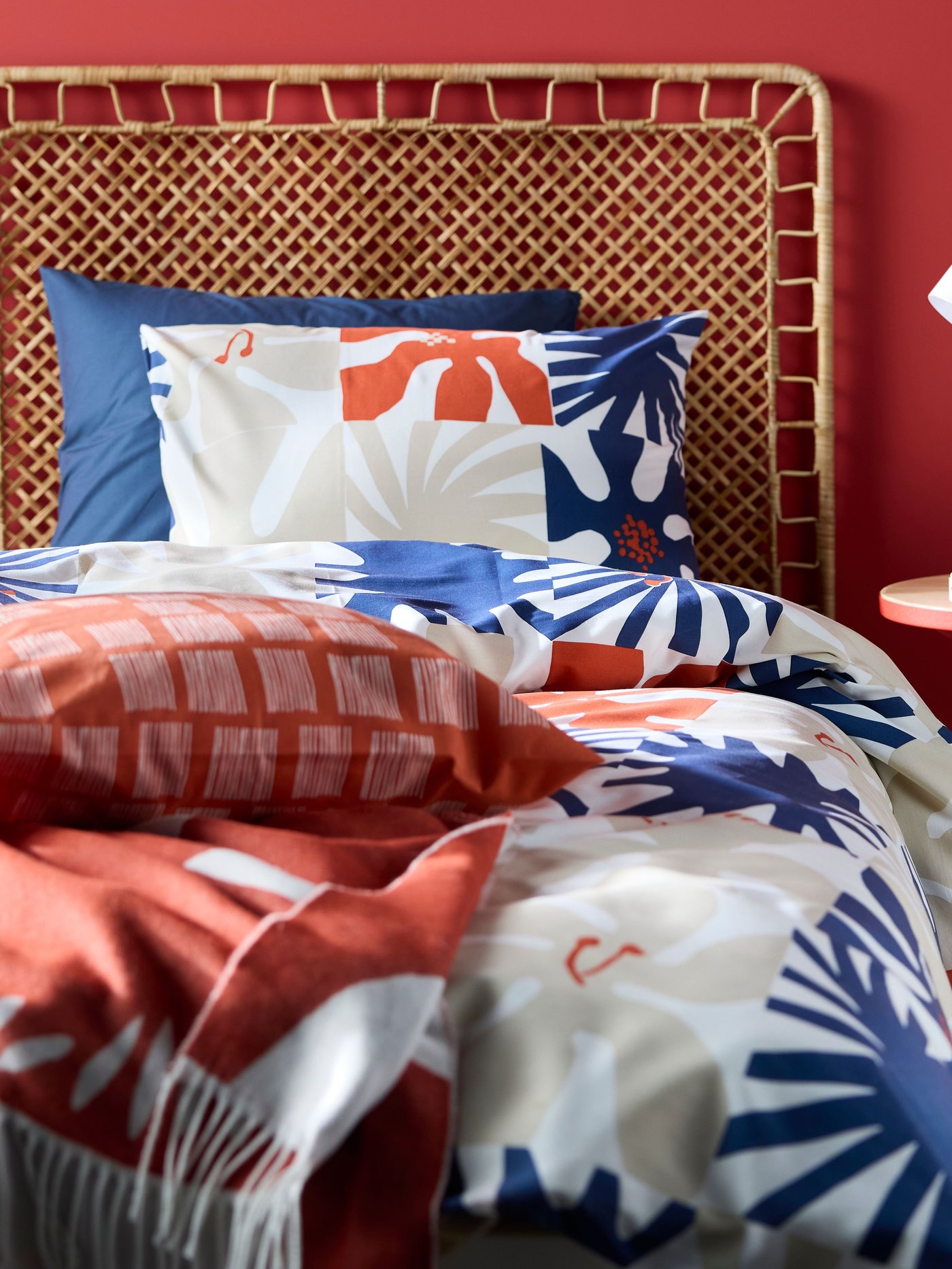 Una lampada da tavolo bianca appoggiata su un tavolino rosso accanto a un letto con testiera in rattan e biancheria da letto HYLSNEJLIKA.