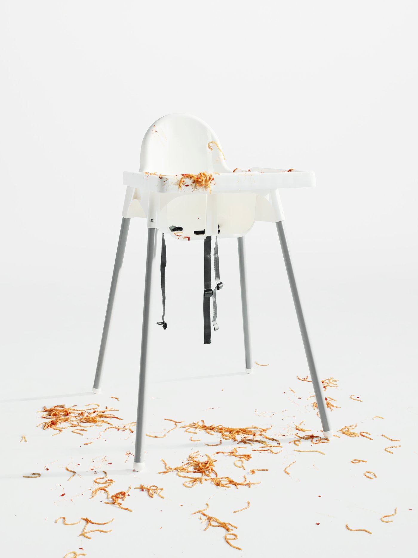Prázdna detská stolička s podnosom v bielom priestore. Zvyšky chaotického jedla zo špagiet pokrývajú detskú stoličku a podlahu.