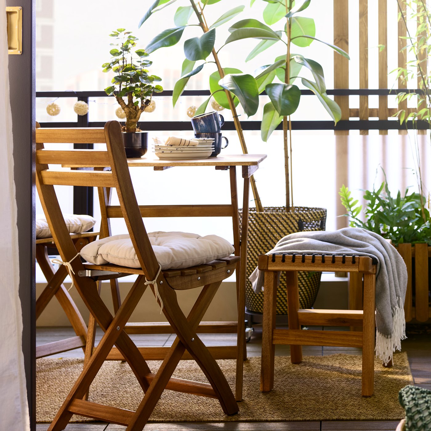 A light brown stained acacia ASKHOLMEN table and foldable chair face the view in a balcony with plants and natural materials.