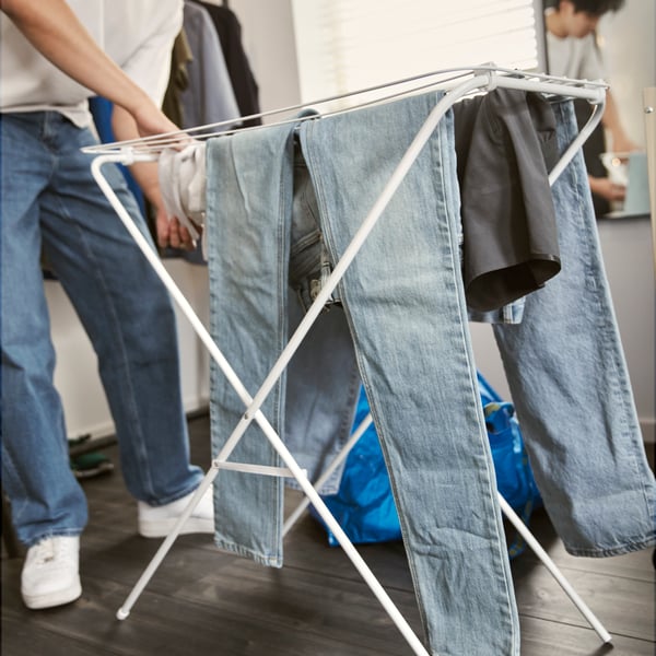 A person wearing jeans is interacting with a JÄLL drying rack in white holding blue jeans and other clothes.