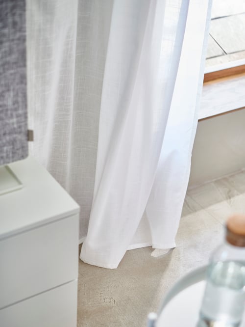 The bottom of a sheer white SILVERLÖNN curtain touching the floor, drawn at the side of a floor-to-ceiling window.