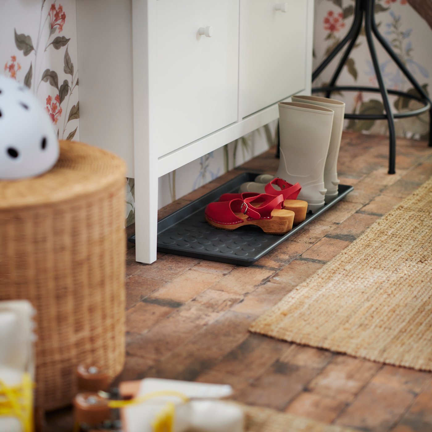 En svart BAGGMUCK skobricka på golvet under ett vitt GULLABERG skoskåp med ett par skor och stövlar ovanpå.