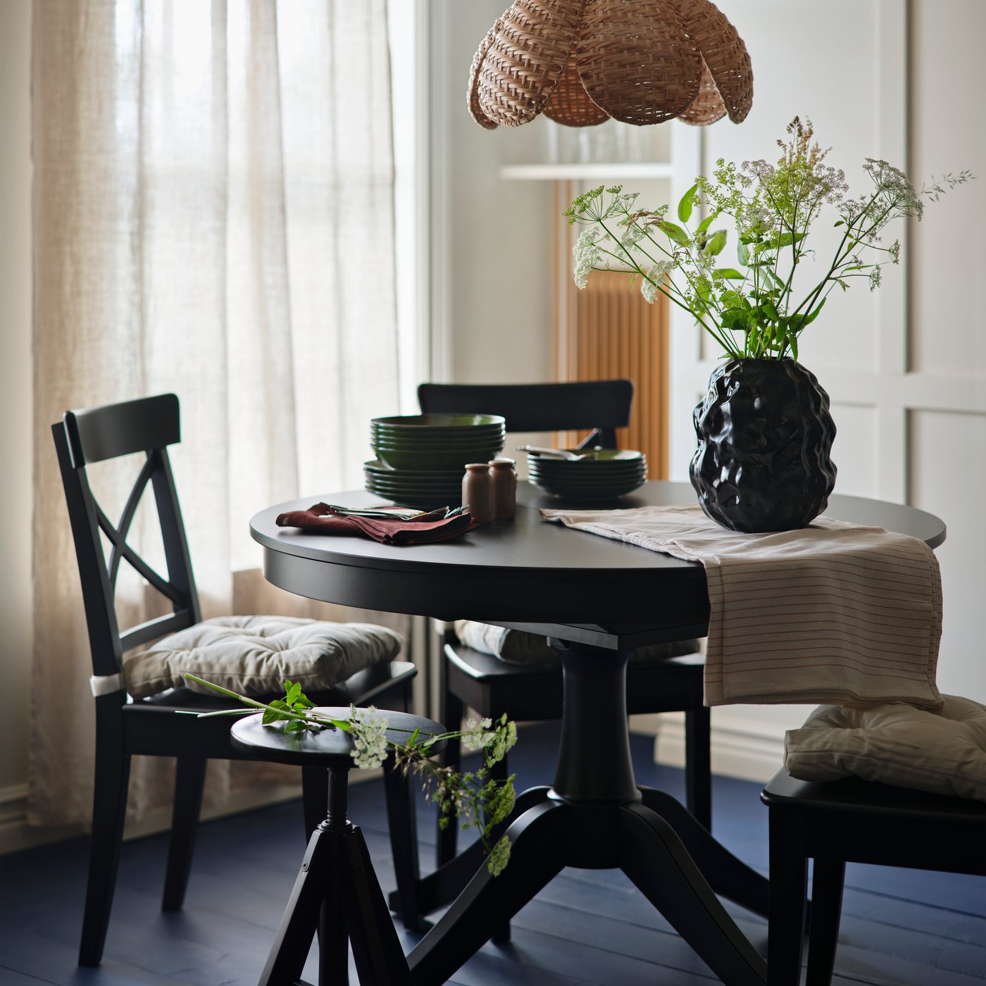 Ett svart utdragbart ROSENTORP-bord är dukat med stolar, porslin och en blomvas i en ljus matsal.