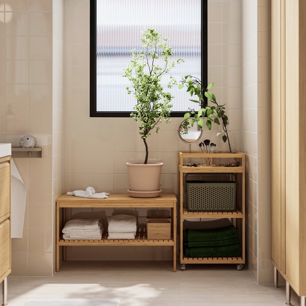A bathroom nook has a bamboo RÅGRUND bench with a potted plant on top and a slatted shelf below with neatly folded towels.