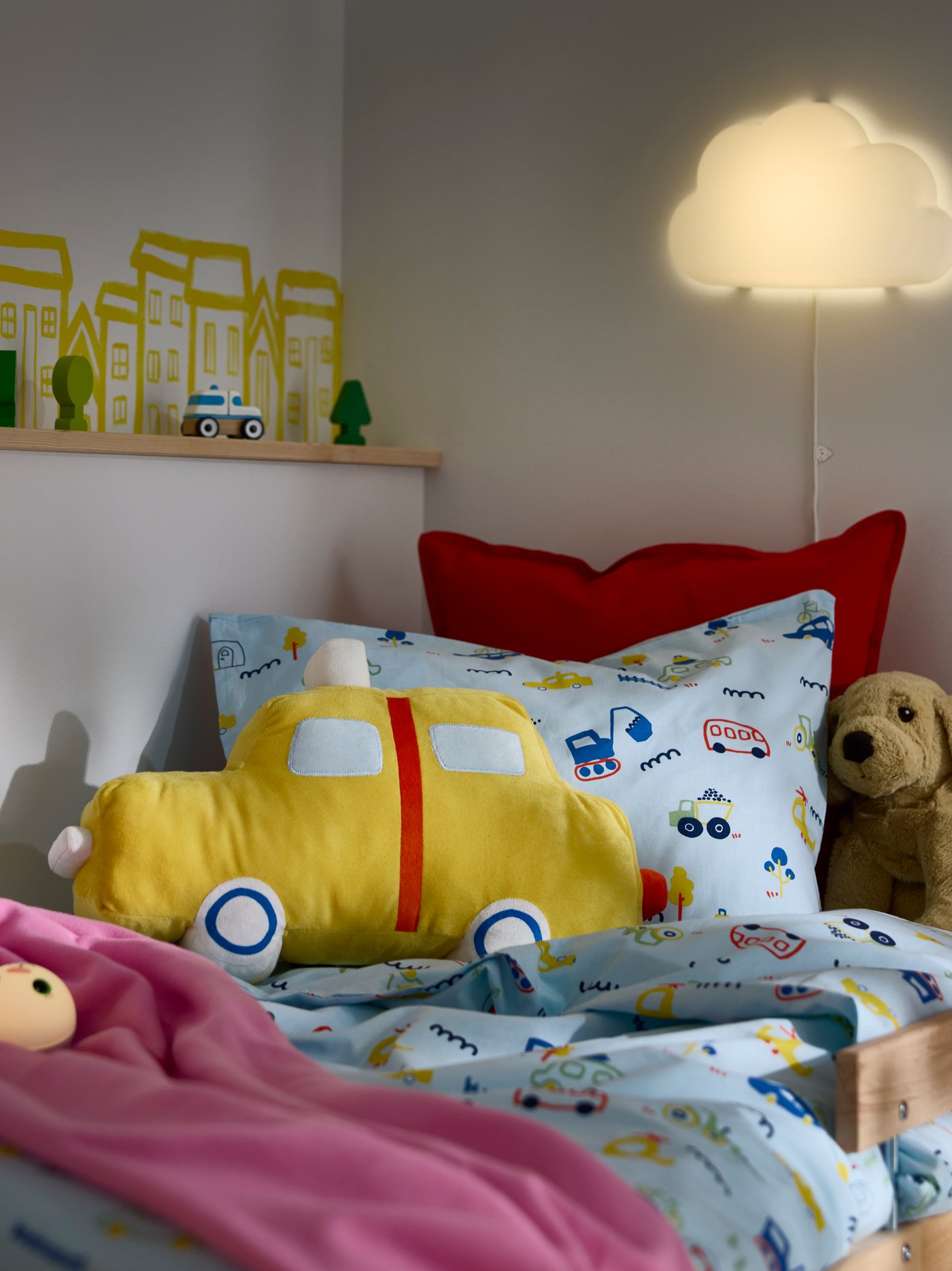 A children’s bed with the BARNDRÖM cushion shaped like a yellow car placed on patterned bedding with toys nearby.
