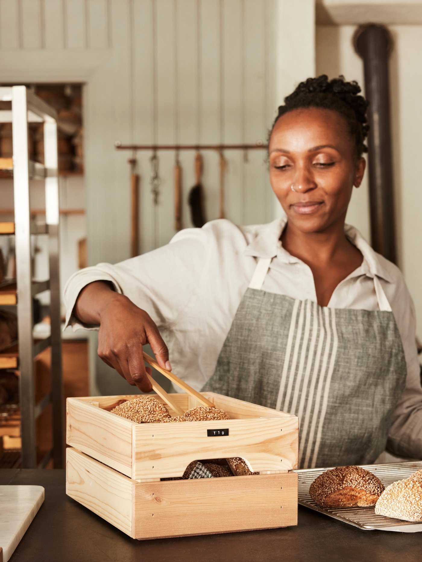 A woman wearing an apron in a café picks fresh bread from a KNAGGLIG crate on the counter.