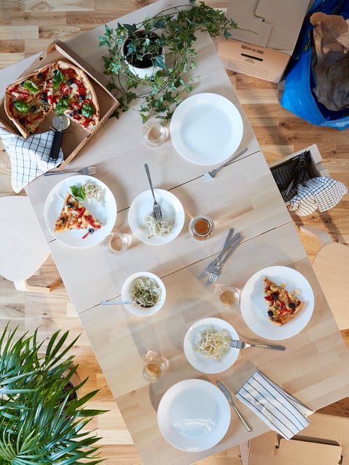 Wooden dining table with white OFTAST plates and bowls and pizza in a box with plants, stools and moving boxes and bags.