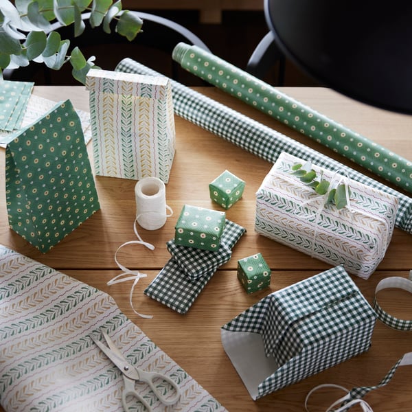 des cadeaux de plusieurs tailles et formes emballés dans des paquets cadeaux aux couleurs nuances de vert et de blanc à côté de ruban blanc et de rouleaux de papier cadeaux