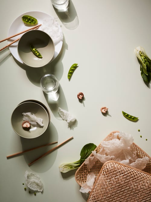 Une présentation artistique incluant des bols verts, une assiette blanche, des baguettes chinoises, une boîte en bambou, des haricots verts dans une cosse et d’autres aliments.