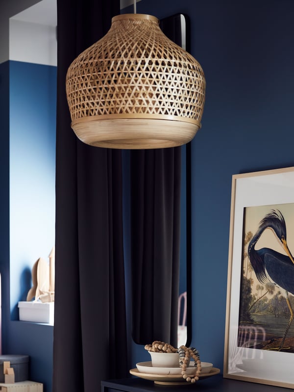 A handmade bamboo MISTERHULT pendant lamp hangs in a room with dark blue walls and dark curtains near a framed picture.
