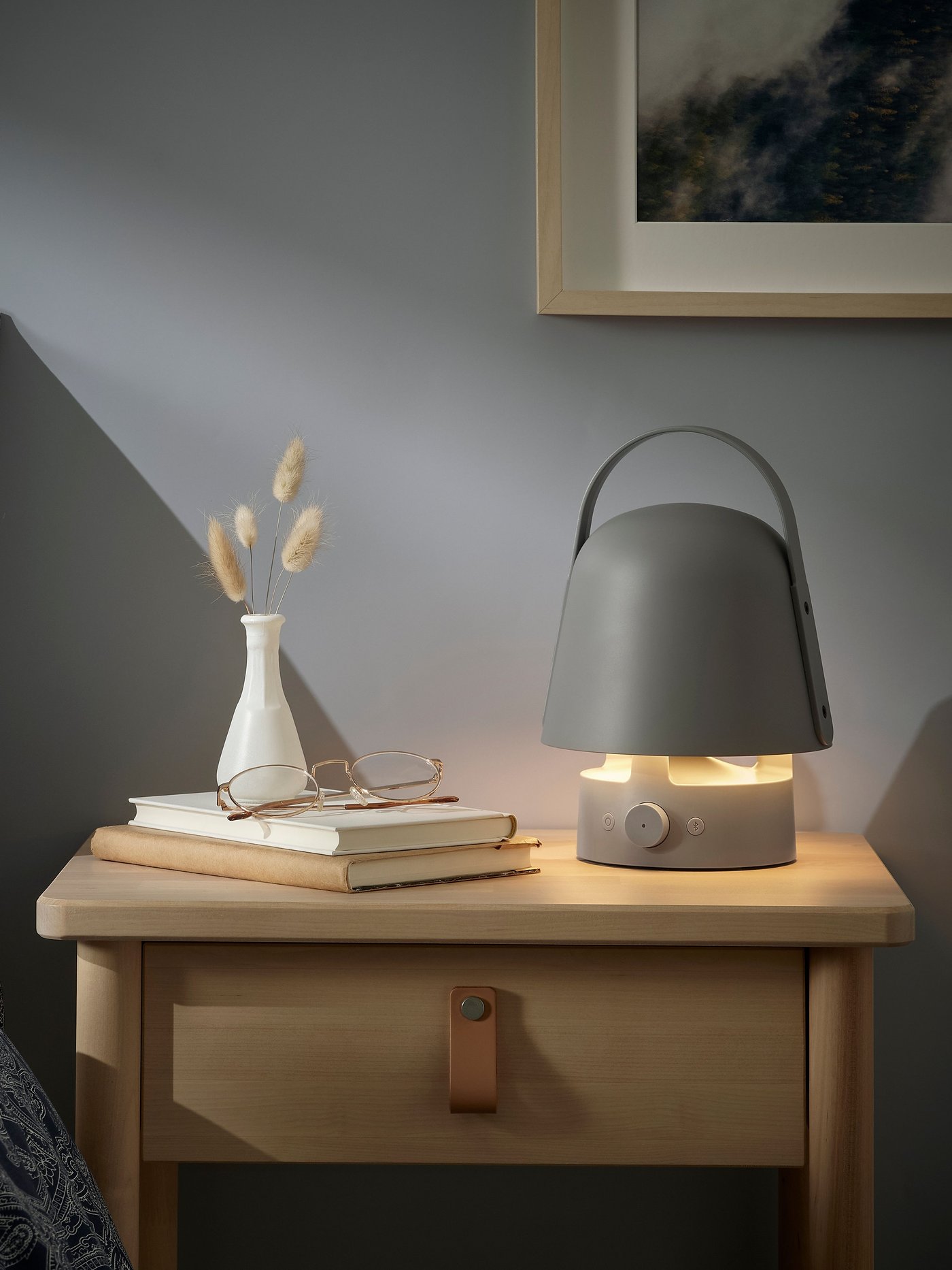 A mushroom-shaped, grey VAPPEBY Bluetooth outdoor speaker lamp with a handle is lit and sitting by books on a bedside table.