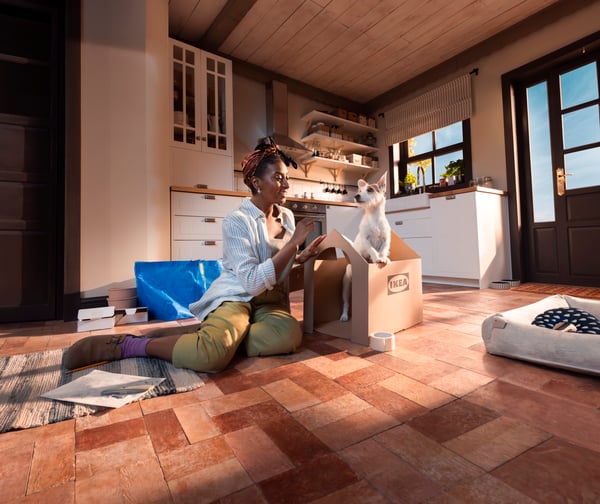 Dans une cuisine moderne, une personne portant une chemise rayée et un pantalon vert est assise par terre et construit une niche pour son chien à partir d’un carton IKEA, le chien se trouvant au milieu du carton. La cuisine est dotée d’armoires en bois blanc et d’électroménagers en acier inoxydable.