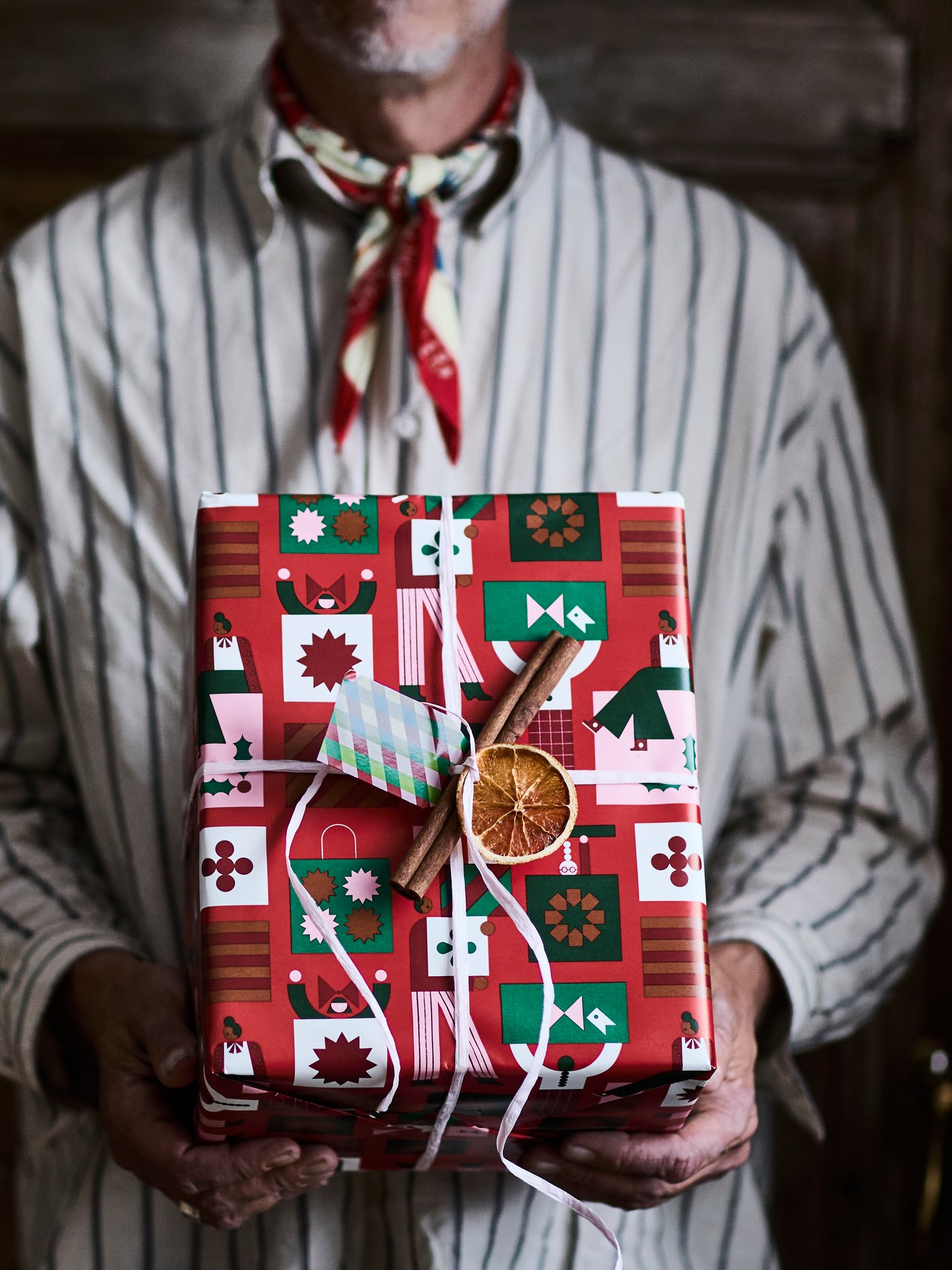 A person with a striped shirt is holding a present wrapped in VINTERFINT gift wrapper and adorned with a dried fruit.