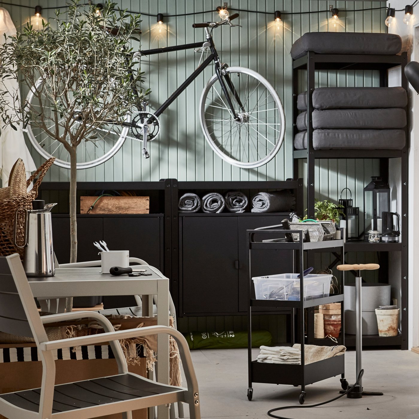 A garage with bike, a lit lighting chain, a wall of storage, a seating area and a black NISSAFORS trolley holding tools.