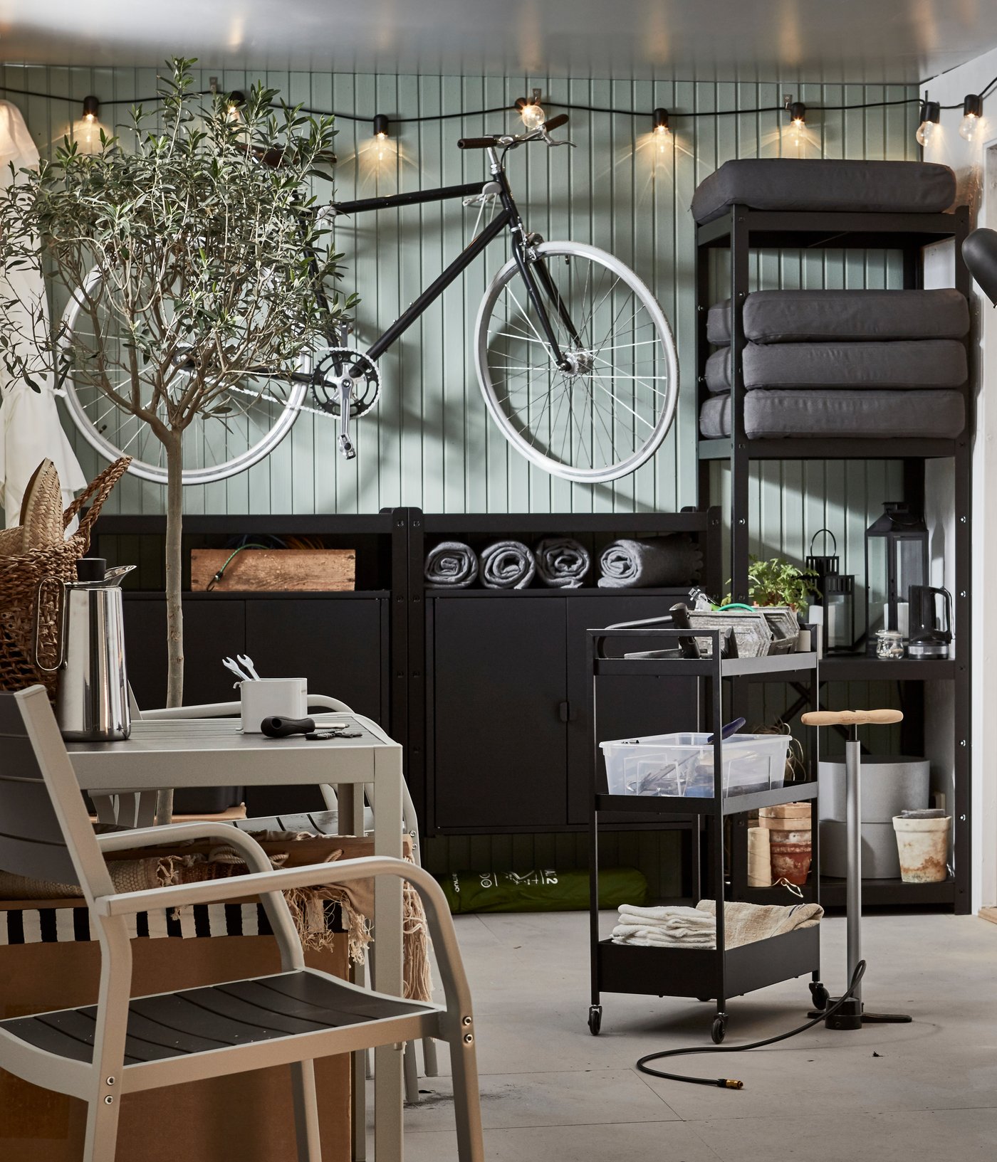 A garage with bike, a lit lighting chain, a wall of storage, a seating area and a black NISSAFORS trolley holding tools.