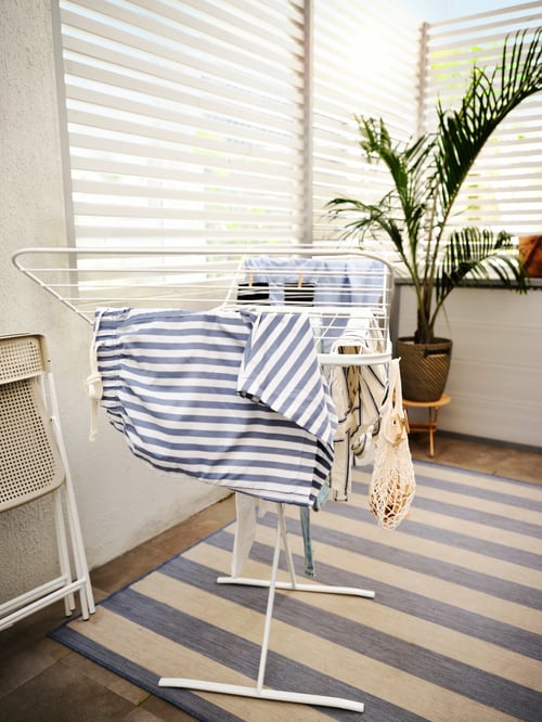 A white MULIG drying rack with clothes on a blue striped VRENSTED rug, a folded TORPARÖ chair and a big plant.