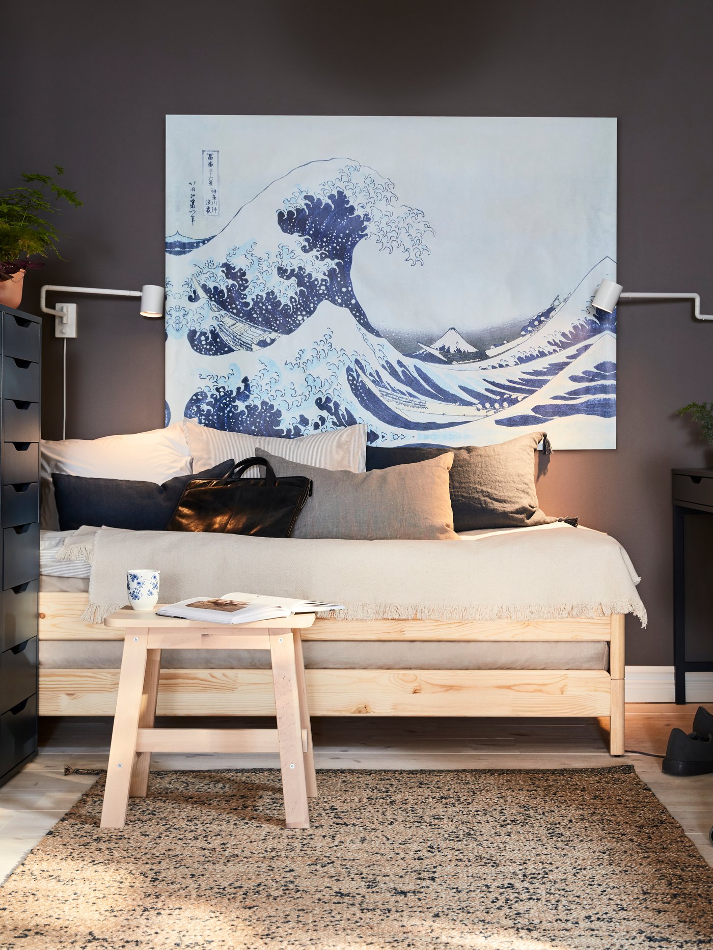 Postel z borovice a březová stolička s knihou a hrnkem vpředu, velký obraz s vlnou a dvě nástěnné lampy.