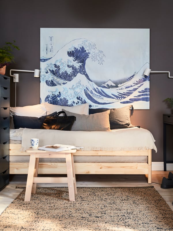 A bed in pine and a birch stool with a book and a mug in front, a big painting with a wave and two wall-mounted lamps.