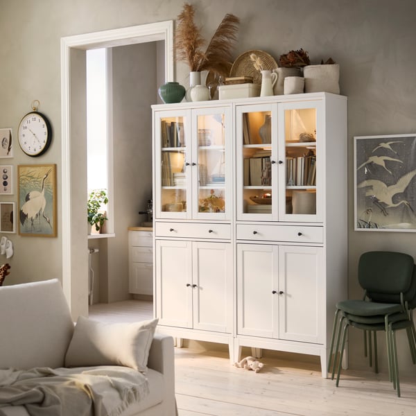 Two white IDANÄS high cabinets with glass doors and 1 drawer, placed side by side, have books and keepsakes stored inside.