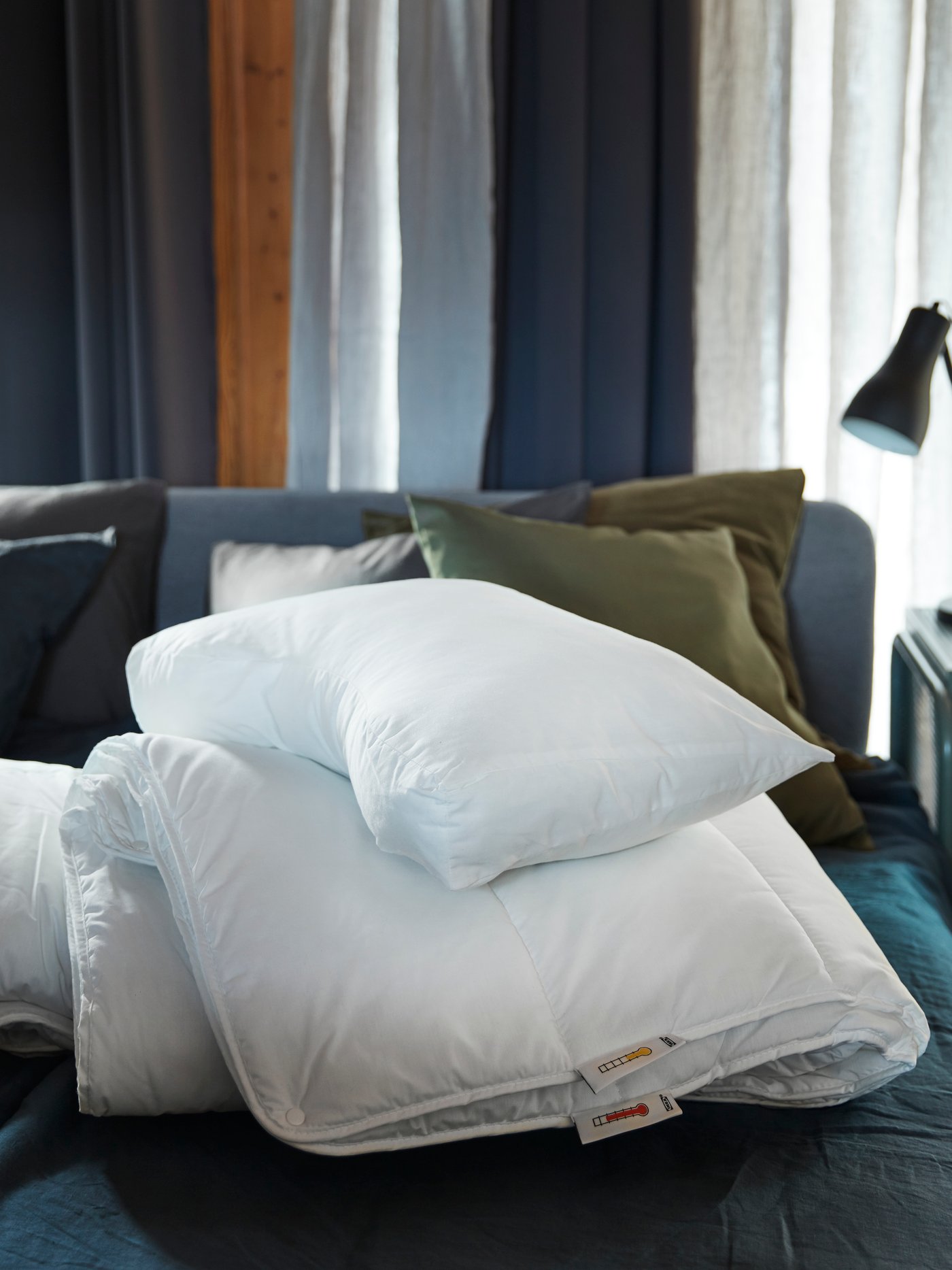 Una cama con ropa de cama azul, un edredón SMÅSPORRE blanco, una almohada ergonómica SKOGSLÖK blanca y cojines verdes de fondo.