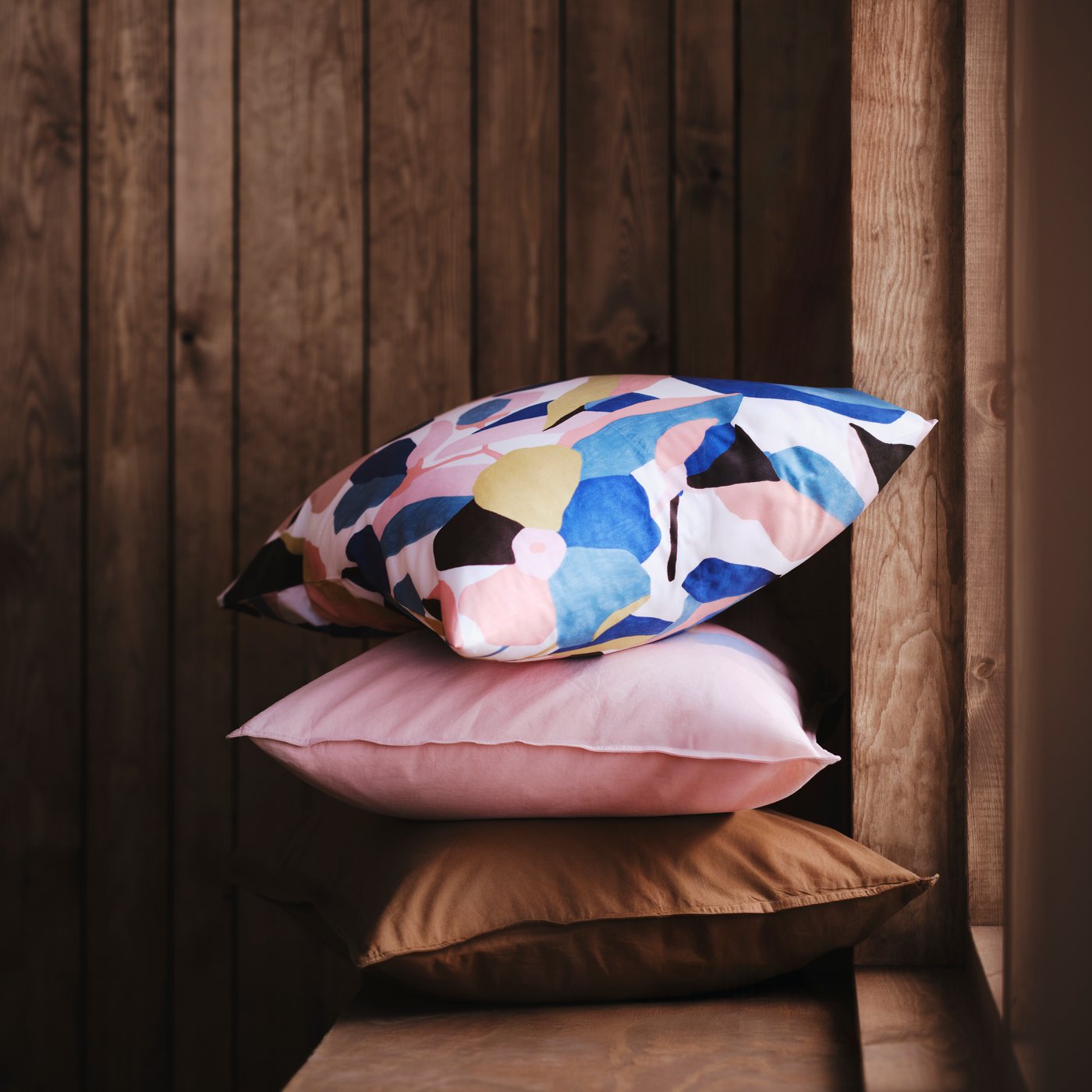 A stack of three pillows, one in a ÄNGSLILJA pillowcase in light pink, sit on a wooden bench against a wood-paneled wall.