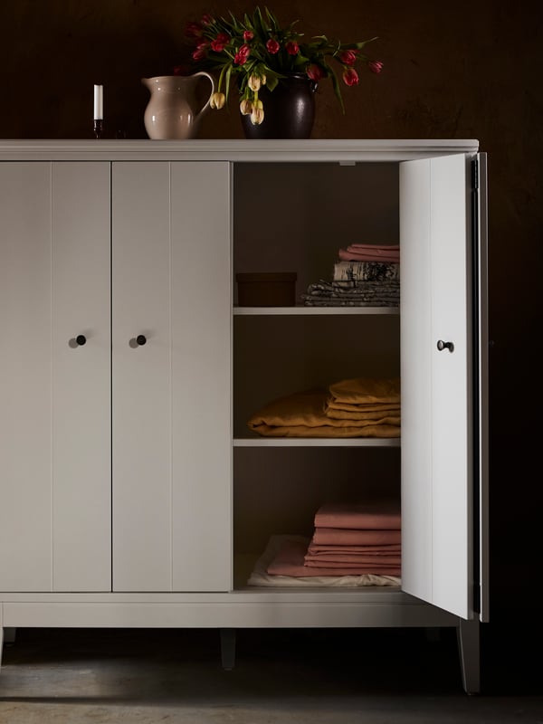 An IDANÄS cabinet in grey with one bi-fold door open showing folded textiles on the shelves. Two decorative jugs are on top.