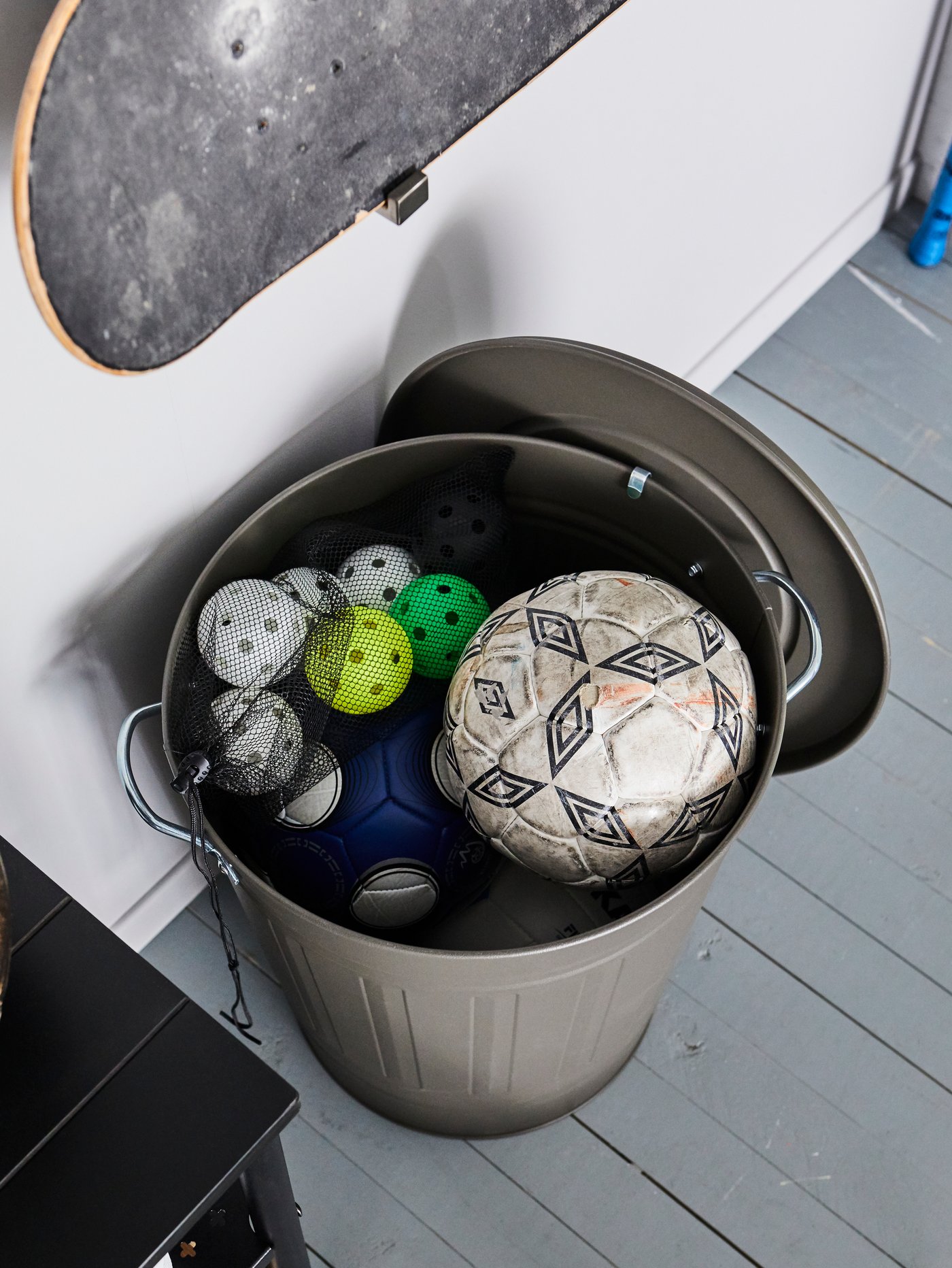 Un basurero gris KNODD con la tapa abierta que muestra pelotas de diferentes tamaños, incluidas dos pelotas de fútbol. Hay una skate en la pared de arriba.