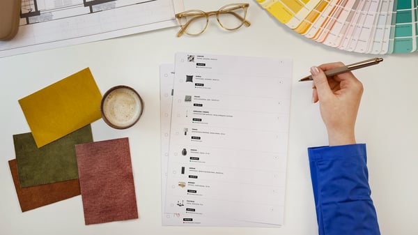 Projektantka wnętrz IKEA prezentująca kartki z wydrukiem listy produktów wykorzystanych w zaprojektowanym pomieszczeniu. Obok na blacie leży wzornik kolorów i próbki materiałów.