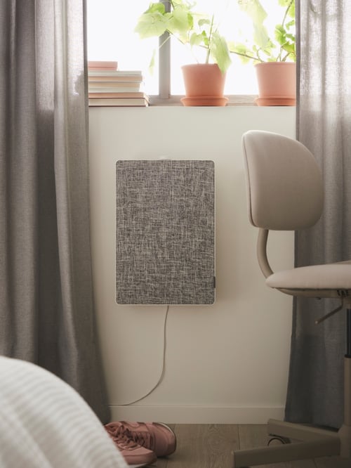 Un purificateur d’air FÖRNUFTIG sur un mur, sous une fenêtre, avec des plantes et des livres posés sur le rebord de la fenêtre, près d’une chaise pivotante.