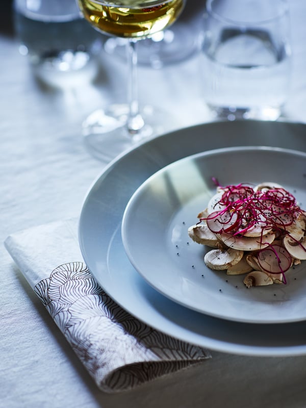 A dining table set with IKEA 365+ white plate with a smaller one on top, next to a patterned napkin, a glass of white wine.