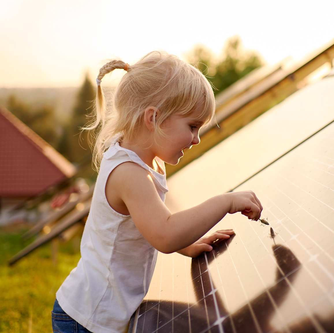 Ett litet barn med blont hår rör vid en solpanel utomhus, med grönska och ett hustak i bakgrunden.