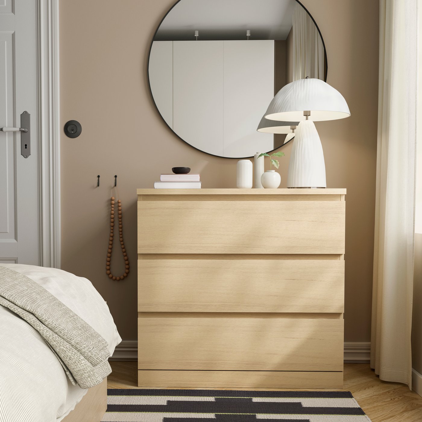 Cassettiera MALM a tre cassetti in legno chiaro, posizionata sotto uno specchio rotondo. Sopra la cassettiera si trovano una lampada bianca, vasi decorativi e libri. Sul pavimento, davanti alla cassettiera, è presente un tappeto con motivi geometrici in bianco e nero.