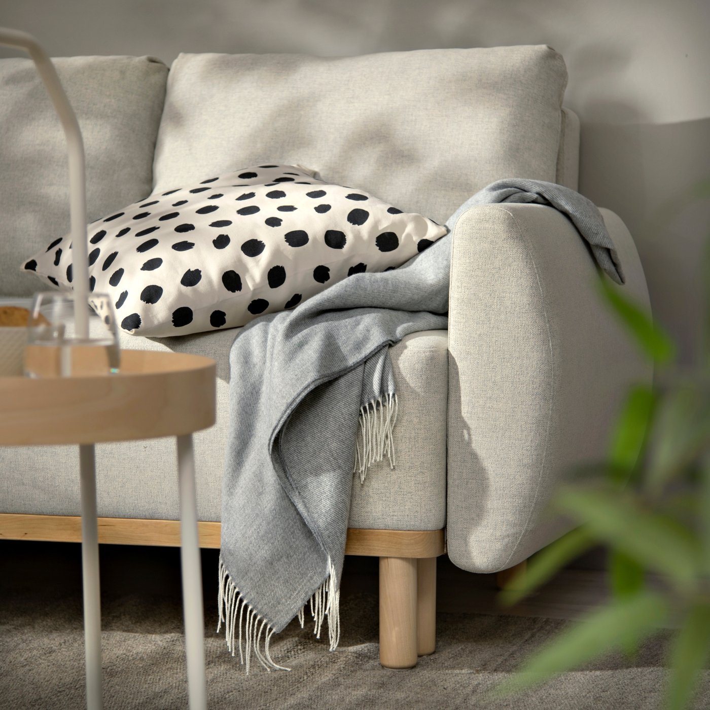 Svijetla sofa s drvenim nogama, na naslonu je bijeli jastuk s crnim točkama i preko naslona prebačena siva deka s resama. Ispred je okrugli drveni stolić, a u kutu vidljiva zelena biljka.