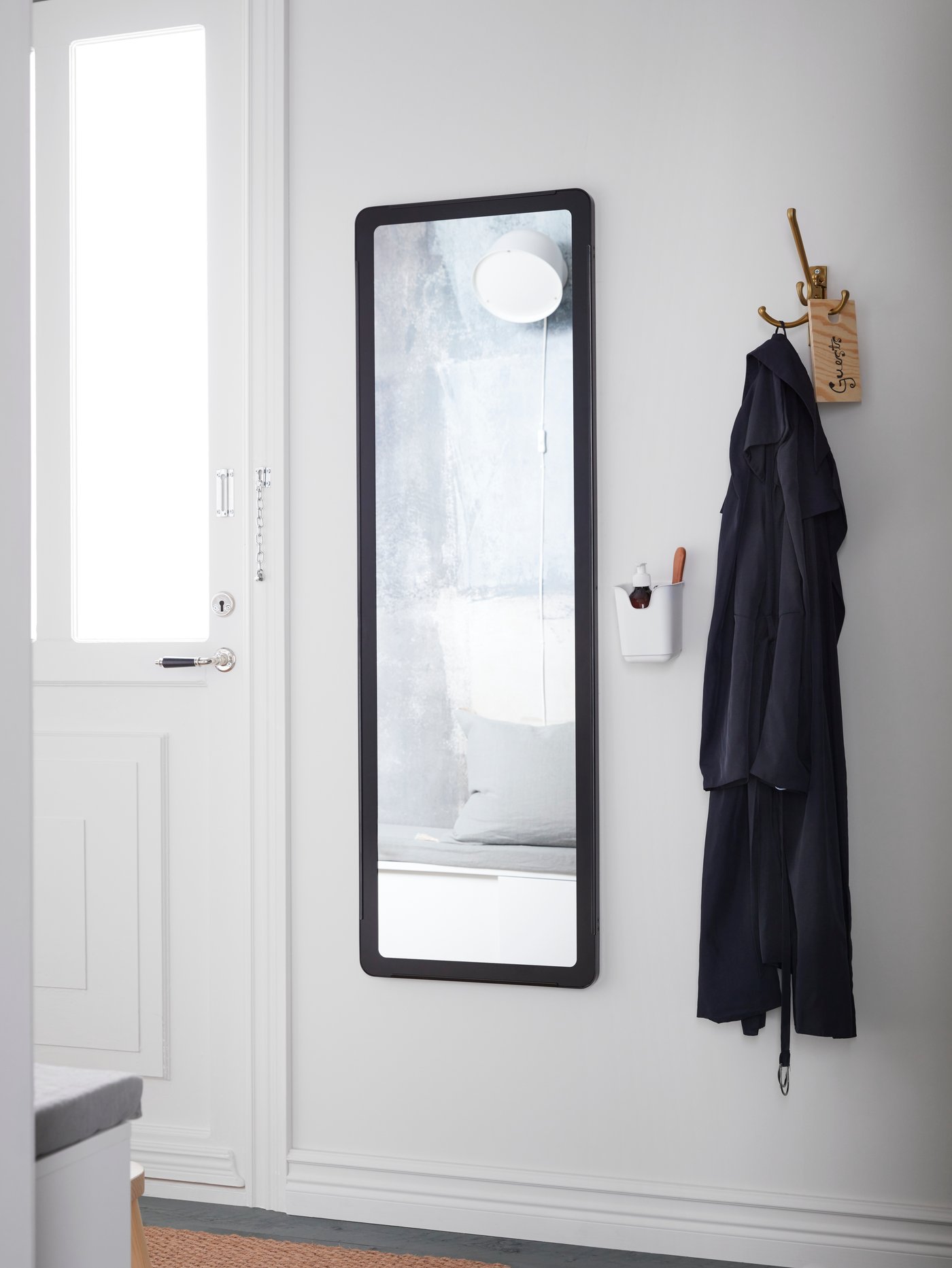 Un mirall GRUA llarg amb un marc negre entre una porta blanca i un ganxo triple, on hi ha un abric penjat.