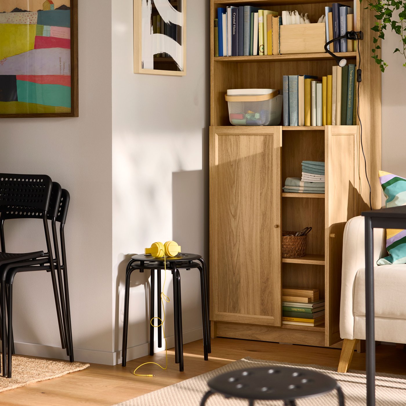 A sunlit room with colourful wall art, stacked chairs and stools and an oak-effect BILLY/OXBERG bookcase in a corner.