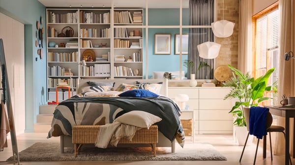 An off- white TONSTAD bed covered in MYGGLASVINGE bed linen in an airy bedroom in front of three white BILLY bookshelves.