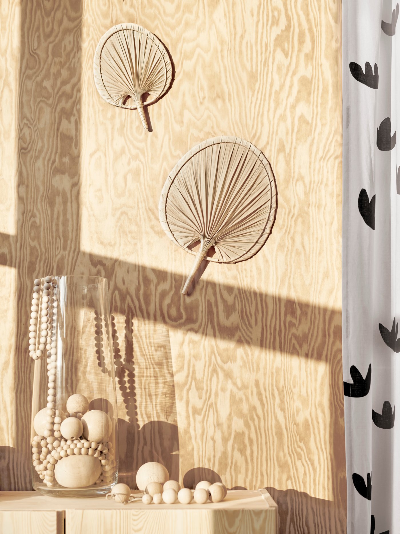 Dormitorio con muebles de madera, objetos personales, dos adornos con hojas de palmera, un jarrón de vidrio, un par de cortinas y joyas.