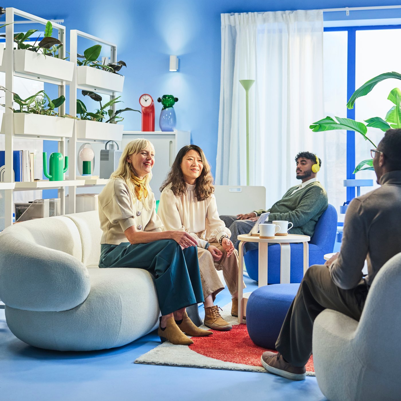 Four people seated in a cozy lounge with plants and soft lighting, talking around a small table with mugs and a tablet.