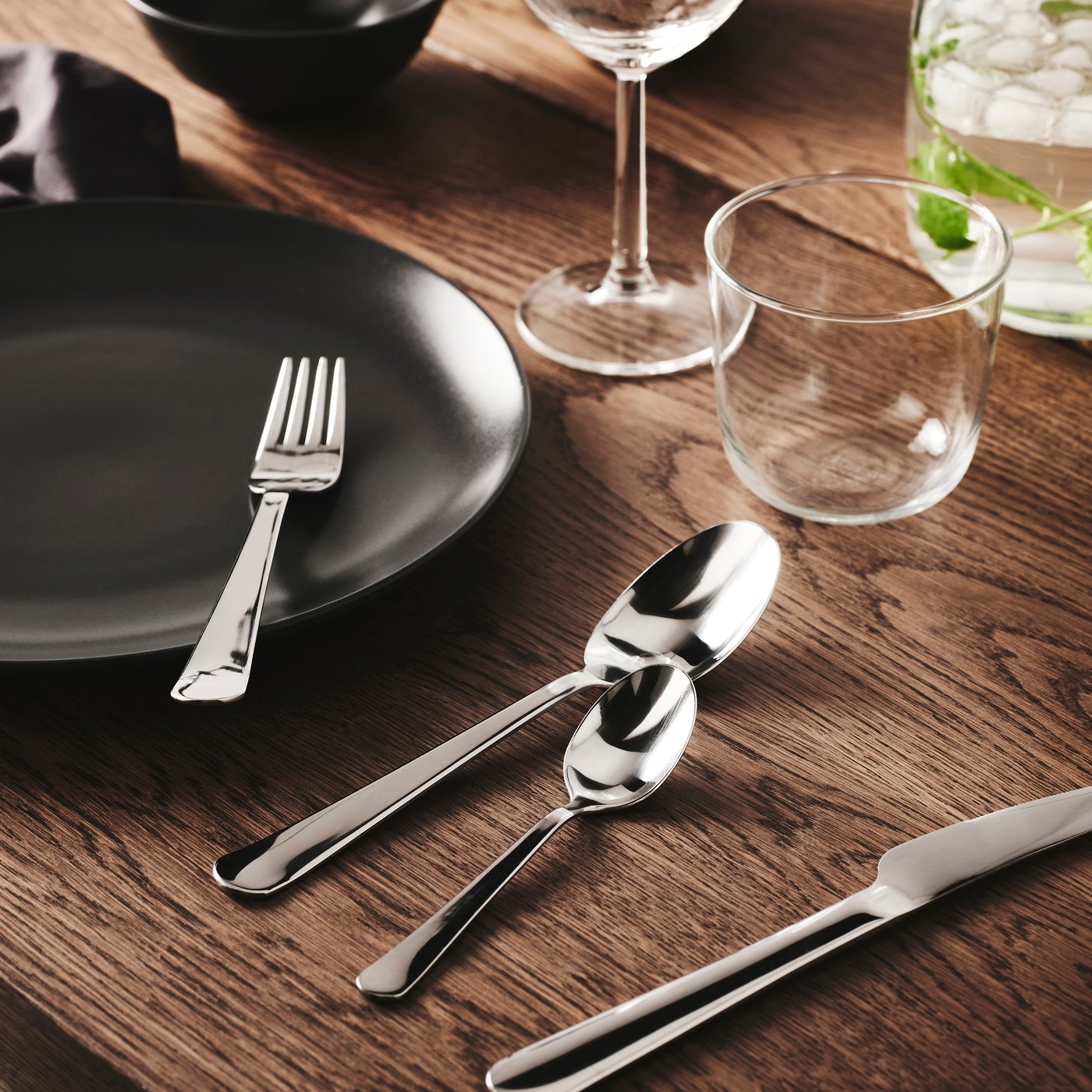 Set table with black plate, glassware and shiny silver cutlery on wooden surface, arranged neatly for a modern dining setting. Stylish decor add warmth