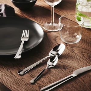 Table dressée avec assiette noire, verres et couverts brillants sur surface en bois, soigneusement arrangés pour un repas moderne à l’ambiance chaleureuse.