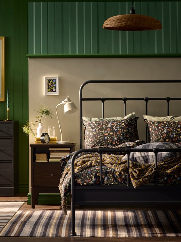 Letto STJÄRNÖ dalla struttura in acciaio color antracite, ricoperto da biancheria floreale, appoggiato ad una parete verde e bianca, con accanto un comodino nero. 