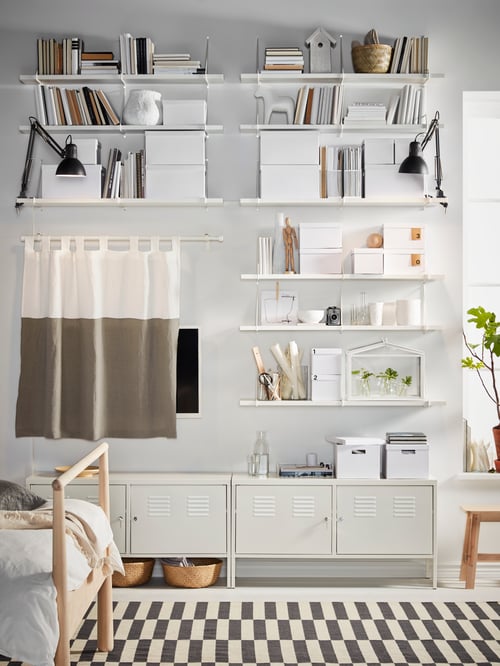Eine Wand mit BERGSHULT/PERSHULT Wandregalen voller Bücher und Boxen und einem Vorhang, hinter dem ein Fernseher und zwei Schränke versteckt sind.