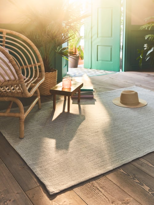 A plank floor has a rectangular, flatwoven TIPHEDE rug in natural/off-white cotton under a rattan armchair and small stool.