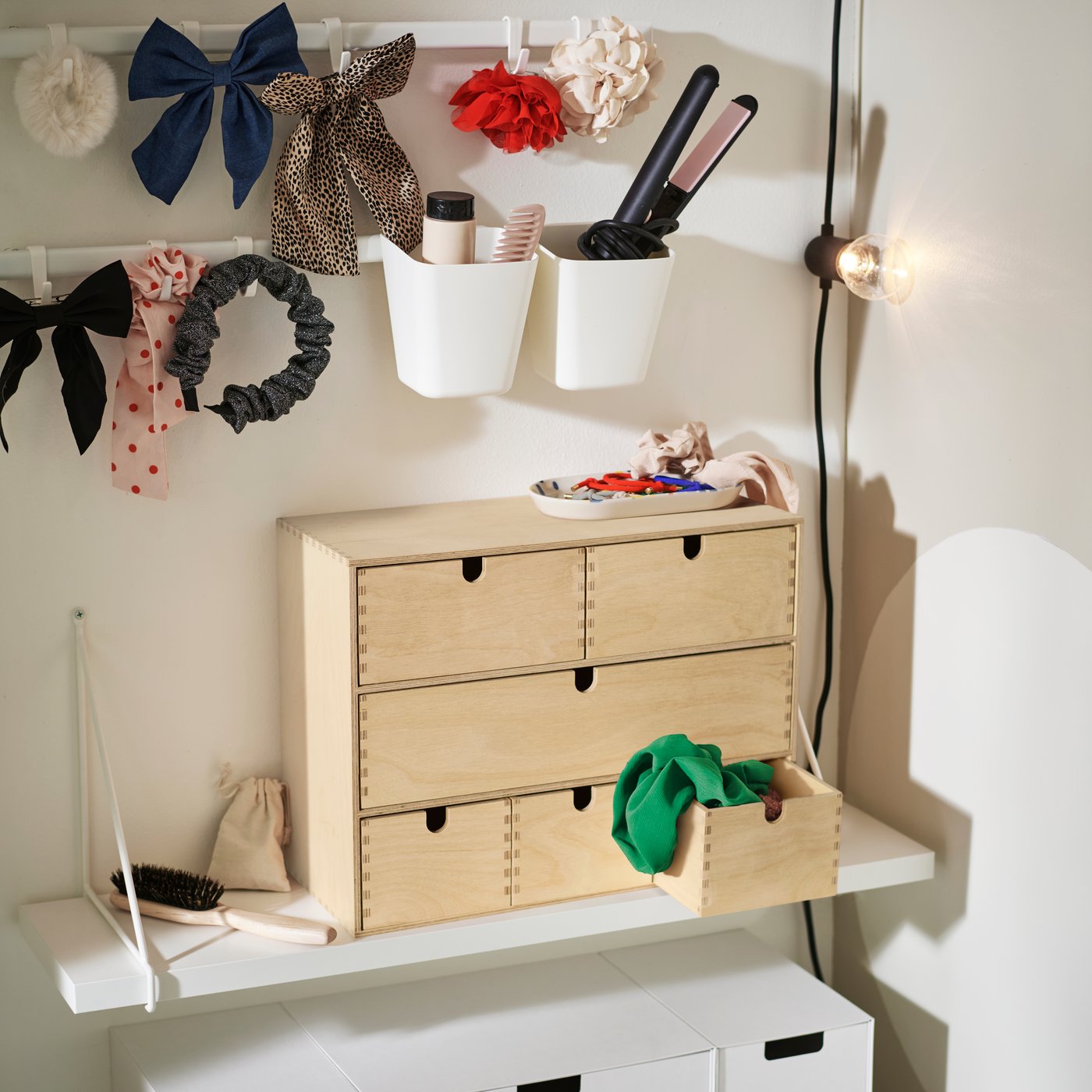 Na bílé polici BERGSHULT je umístěna mini komoda MOPPE z borového dřeva, ve které jsou uloženy vlasové doplňky, a nad ní jsou dvě háčky.